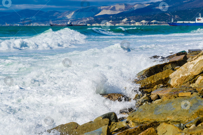 Скачать Курорт Кабардинка. Большие зеленые штормовые волны в белой пене накатываются на каменистый пляж Кабардинки. На заднем плане - порт Новороссийск и Кавказские горы. Большие камни на берегу. Брызги и пена от волн фотосток Ozero