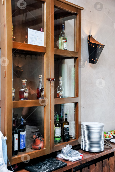 Скачать Деревянный шкафчик с различными бутылками из-под ликера и стопками тарелок в уютной кухне фотосток Ozero