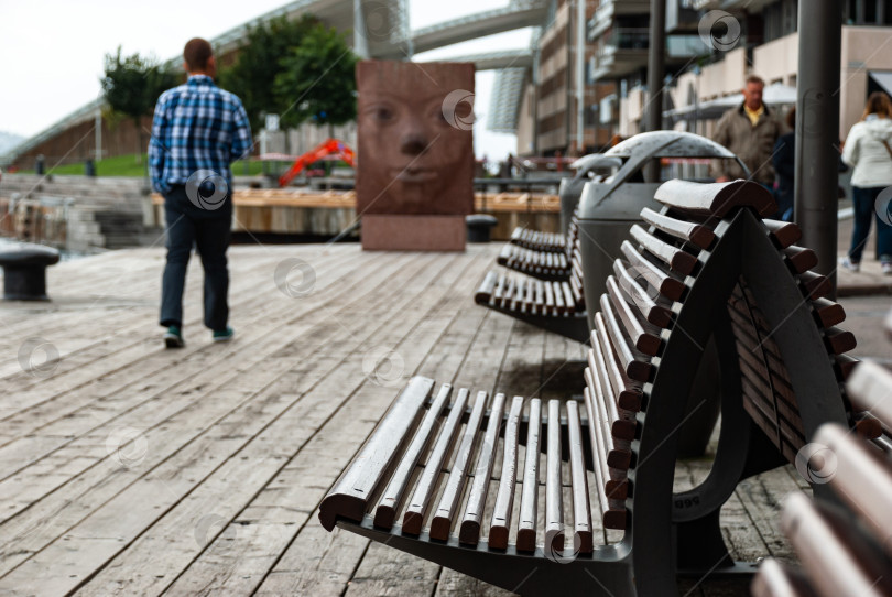 Скачать Сцена на городской набережной с мужчиной, прогуливающимся мимо скульптур и скамеек фотосток Ozero