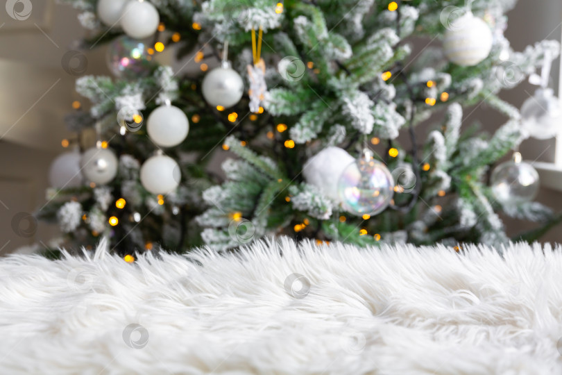 Скачать Рождественский или новогодний фон. Белый меховой коврик на фоне рождественской елки, украшенной горящей гирляндой фотосток Ozero