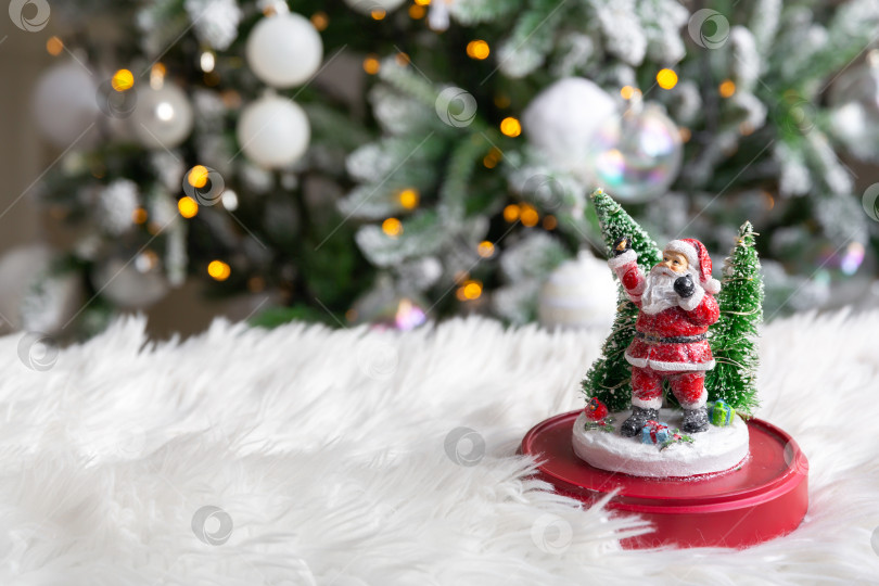 Скачать фигурка Деда Мороза на белом меховом одеяле, на фоне горящей гирлянды рождественской елки. фотосток Ozero