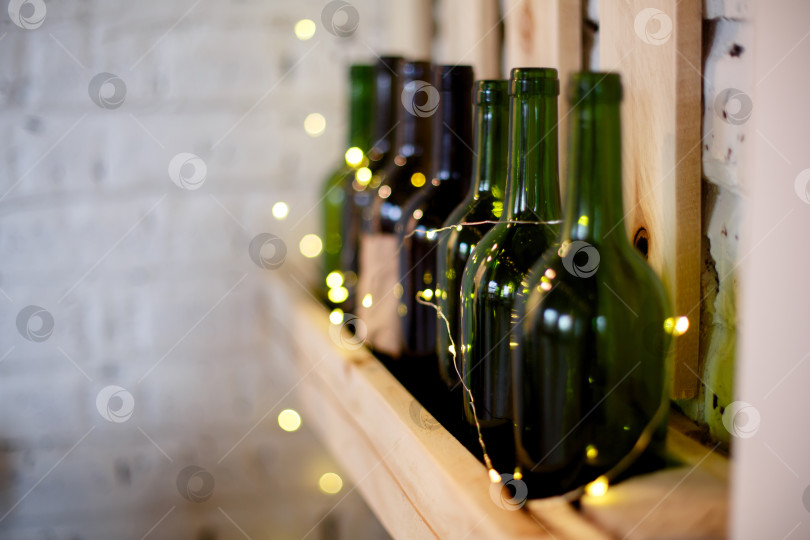 Скачать Желтая рождественская гирлянда и зеленые винные бутылки на деревянной полке фотосток Ozero