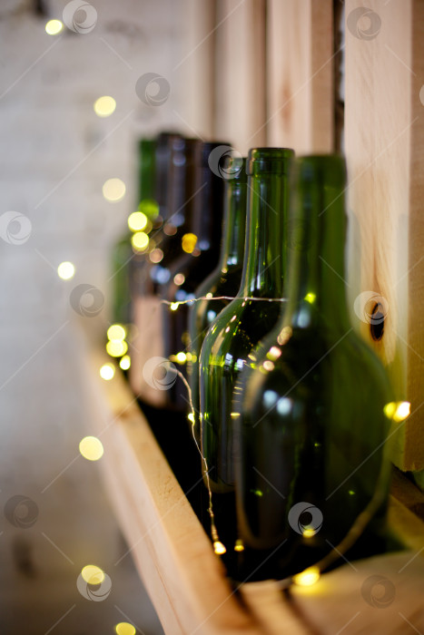 Скачать Желтая рождественская гирлянда и зеленые винные бутылки на деревянной полке фотосток Ozero