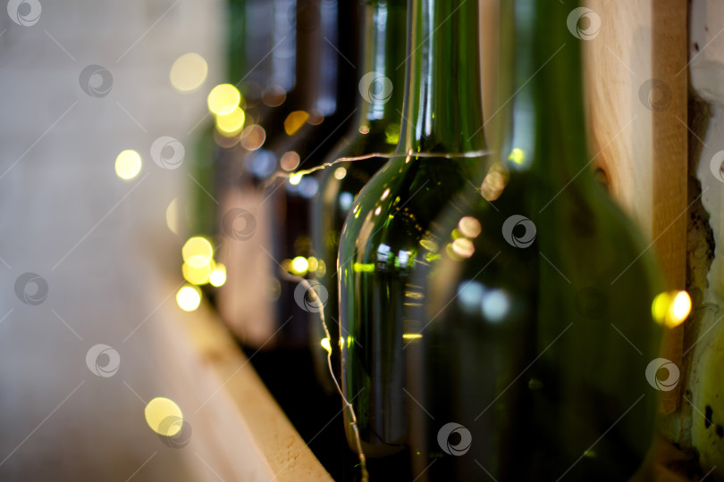 Скачать Желтая рождественская гирлянда и зеленые винные бутылки на деревянной полке фотосток Ozero