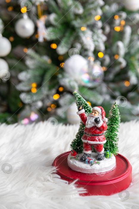 Скачать фигурка Деда Мороза на белом меховом одеяле, на фоне горящей гирлянды рождественской елки. фотосток Ozero