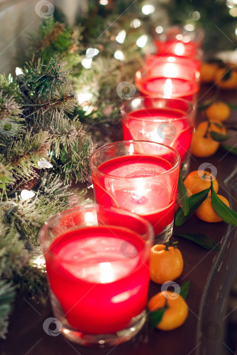 Скачать Свечи в стеклянных подсвечниках стоят на полке вместе со светящейся гирляндой и мандаринами. фотосток Ozero