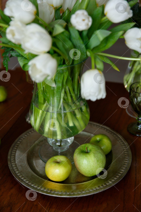 Скачать белые тюльпаны в стеклянной вазе и зеленые яблоки на тарелке на столе фотосток Ozero