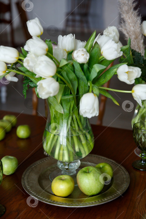 Скачать белые тюльпаны в стеклянной вазе и зеленые яблоки на тарелке на столе фотосток Ozero