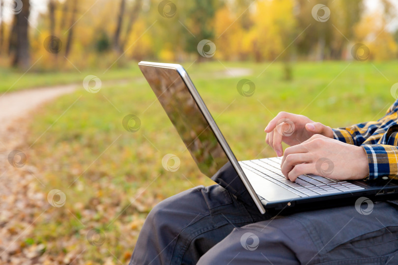 Скачать мужчина сидит в парке и работает за ноутбуком на открытом воздухе. Руки печатают на клавиатуре фотосток Ozero