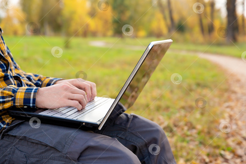 Скачать мужчина сидит в парке и работает за ноутбуком на открытом воздухе. Руки печатают на клавиатуре фотосток Ozero