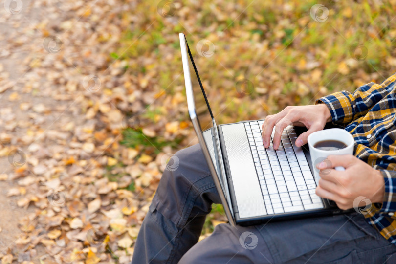 Скачать мужчина сидит в парке и работает на ноутбуке на открытом воздухе. Руки печатают на клавиатуре и держат кружку с кофе фотосток Ozero