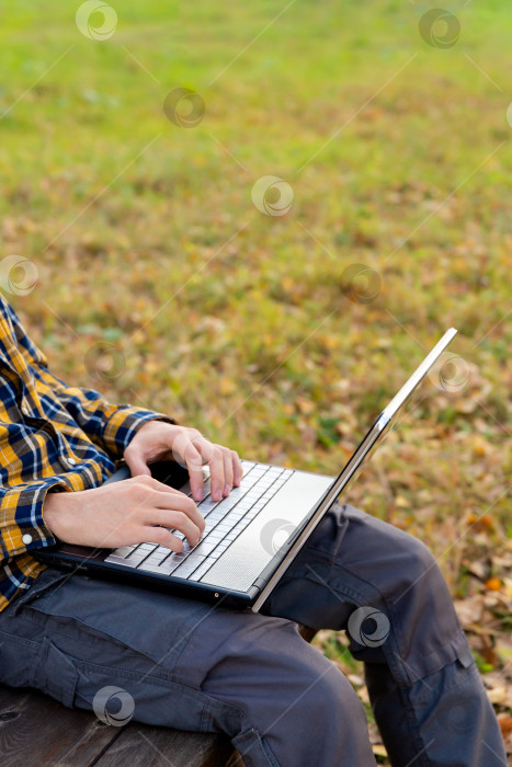 Скачать мужчина сидит в парке и работает за ноутбуком на открытом воздухе. Руки печатают на клавиатуре фотосток Ozero
