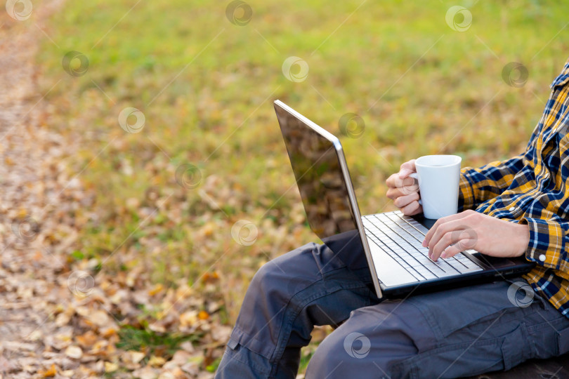 Скачать мужчина сидит в парке и работает на ноутбуке на открытом воздухе. Руки печатают на клавиатуре и держат кружку с кофе фотосток Ozero