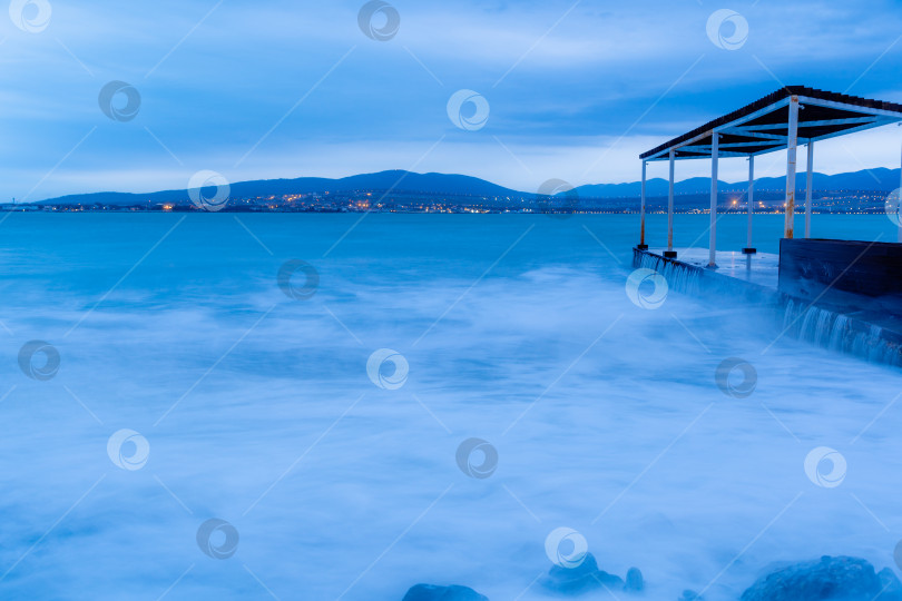 Скачать Шторм в Геленджике на длительной выдержке. Волны в виде тумана набегают на пляж Геленджика. Пирс. Сумерки, закат. Горы и городские огни на горизонте фотосток Ozero