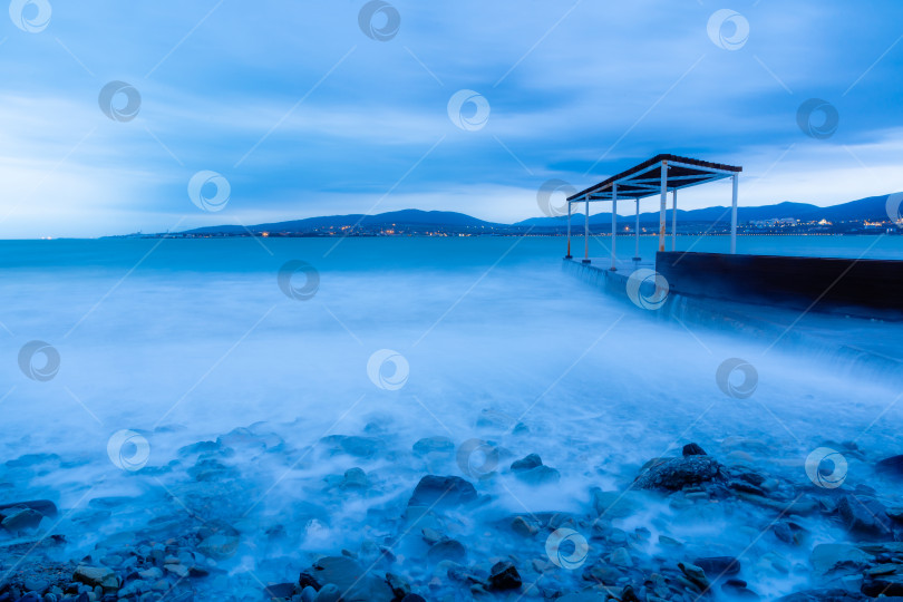 Скачать Шторм в Геленджике на длительной выдержке. Волны в виде тумана набегают на пляж Геленджика. Пирс. Сумерки, закат. Горы и городские огни на горизонте фотосток Ozero