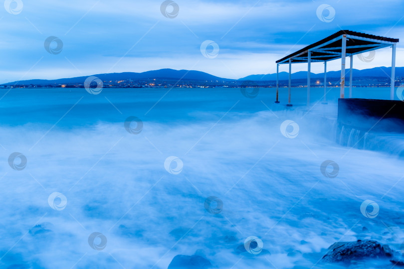 Скачать Шторм в Геленджике на длительной выдержке. Волны в виде тумана набегают на пляж Геленджика. Пирс. Сумерки, закат. Горы и городские огни на горизонте фотосток Ozero