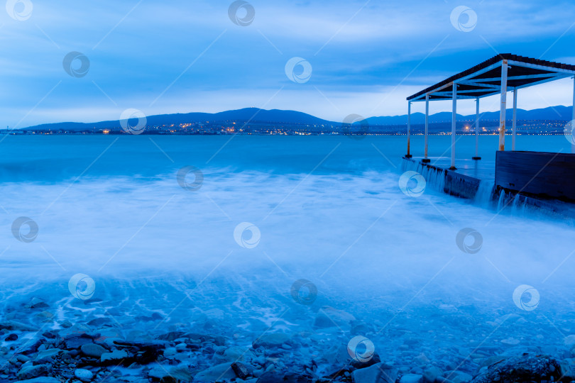 Скачать Шторм в Геленджике на длительной выдержке. Волны в виде тумана набегают на пляж Геленджика. Пирс. Сумерки, закат. Горы и городские огни на горизонте фотосток Ozero