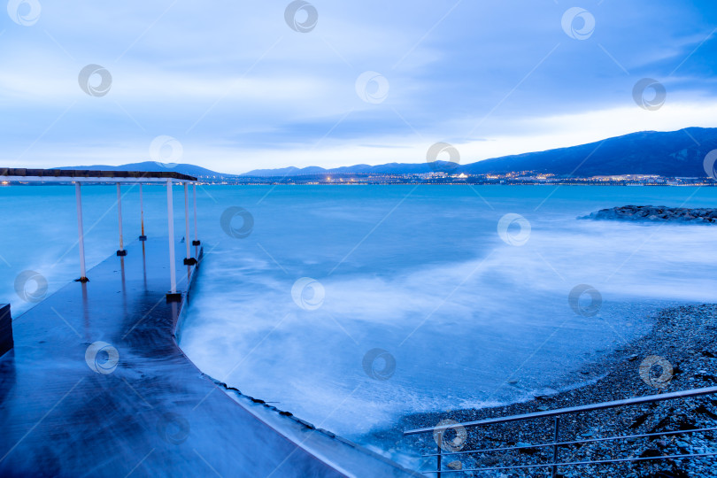 Скачать Шторм в Геленджике на длительной выдержке. Волны в виде тумана набегают на пляж Геленджика. Пирс. Сумерки, закат. Горы и городские огни на горизонте фотосток Ozero