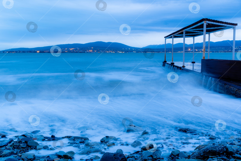 Скачать Шторм в Геленджике на длительной выдержке. Волны в виде тумана набегают на пляж Геленджика. Пирс. Сумерки, закат. Горы и городские огни на горизонте фотосток Ozero