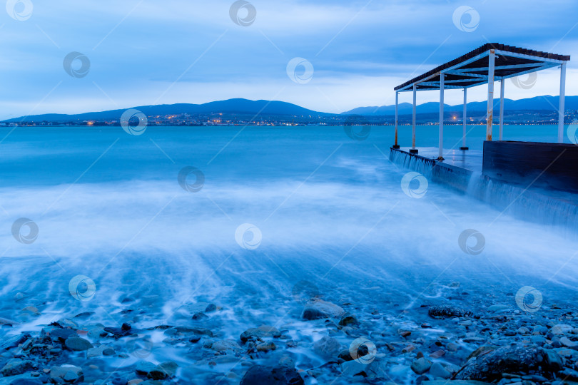 Скачать Шторм в Геленджике на длительной выдержке. Волны в виде тумана набегают на пляж Геленджика. Пирс. Сумерки, закат. Горы и городские огни на горизонте фотосток Ozero