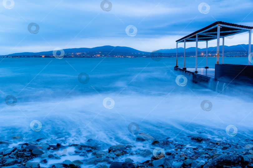 Скачать Шторм в Геленджике на длительной выдержке. Волны в виде тумана набегают на пляж Геленджика. Пирс. Сумерки, закат. Горы и городские огни на горизонте фотосток Ozero