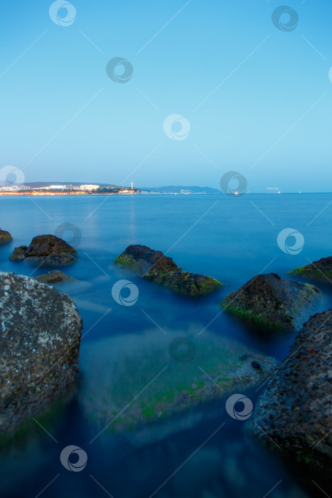 Скачать Геленджикский маяк в вечерних сумерках. Огни маяка отражаются в воде. Маяк стоит на высоком утесе. На переднем плане в воде видны камни, покрытые водорослями фотосток Ozero