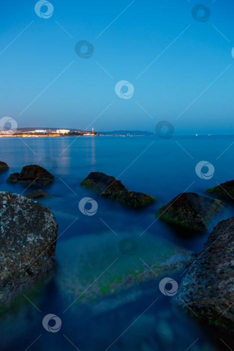 Скачать Геленджикский маяк в вечерних сумерках. Огни маяка отражаются в воде. Маяк стоит на высоком утесе. На переднем плане в воде видны камни, покрытые водорослями фотосток Ozero