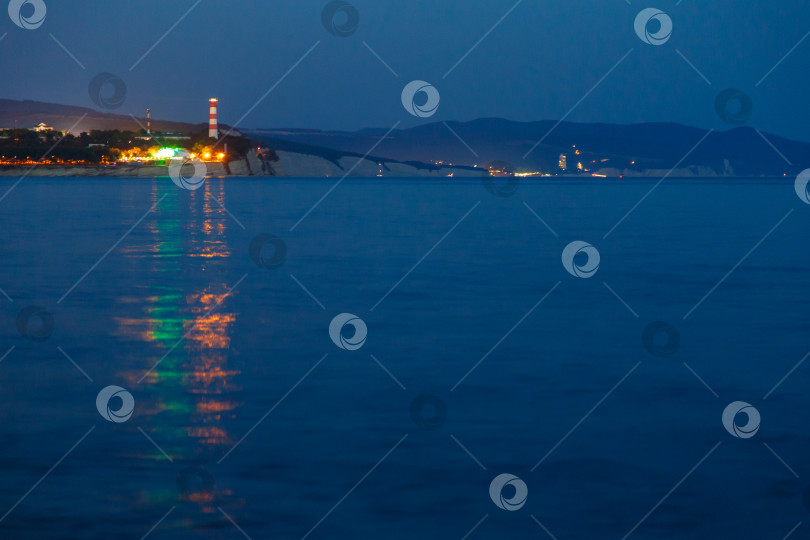 Скачать Геленджикский маяк в вечерних сумерках. Огни маяка отражаются в воде. Маяк стоит на высоком утесе. фотосток Ozero