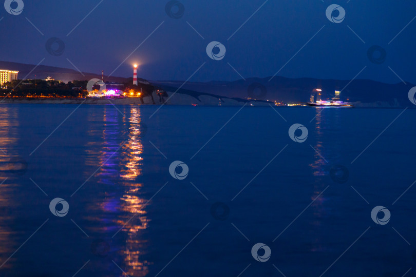 Скачать Геленджикский маяк в вечерних сумерках. Огни маяка отражаются в воде. Маяк стоит на высоком утесе. Мимо маяка проплывает прогулочный катер фотосток Ozero