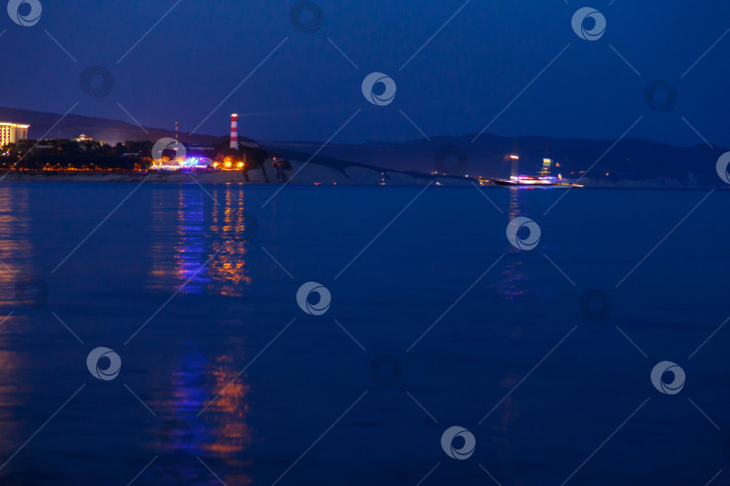 Скачать Геленджикский маяк в вечерних сумерках. Огни маяка отражаются в воде. Маяк стоит на высоком утесе. Мимо маяка проплывает прогулочный катер фотосток Ozero