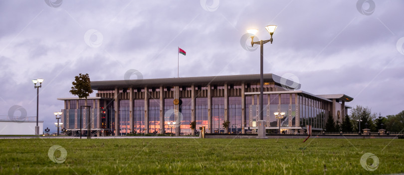 Скачать Современное правительственное здание в сумерках с освещенными окнами и белорусским флагом фотосток Ozero