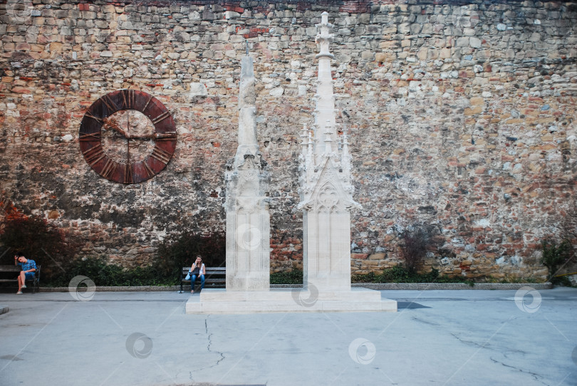 Скачать Готическая скульптура у исторической каменной стены с сидящими людьми в городской обстановке фотосток Ozero