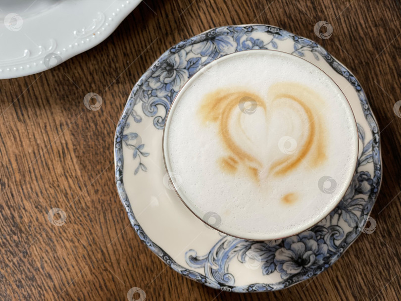 Скачать Капучино с латте-артом в виде сердца в синей чашке с цветочным рисунком на фоне деревянного стола фотосток Ozero
