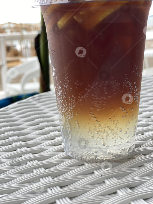 Скачать Холодный чай в прозрачном пластиковом стаканчике с ломтиками лимона и пузырьками на плетеном столике на фоне моря фотосток Ozero