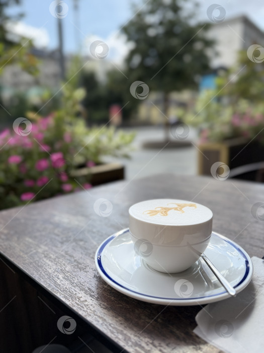 Скачать Чашка капучино с латте-артом на столике кафе на открытом воздухе на фоне городского пейзажа фотосток Ozero