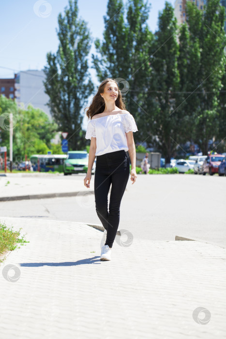 Скачать Молодая красивая брюнетка в джинсах и белой блузке прогуливается по летней улице фотосток Ozero
