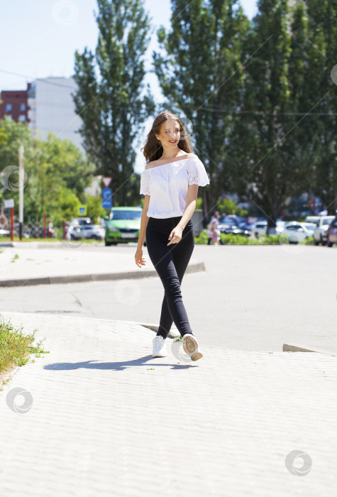 Скачать Молодая красивая брюнетка в джинсах и белой блузке прогуливается по летней улице фотосток Ozero