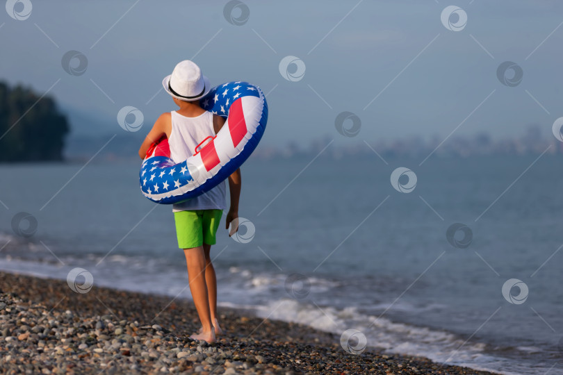 Скачать Летним утром недалеко от берега мальчик прогуливается по пляжу с надувным кольцом в цветах американского флага фотосток Ozero
