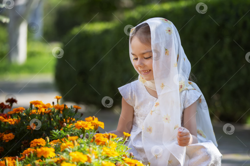 Скачать Молодая девушка в белом шарфе улыбается, ухаживая за оранжевыми цветами календулы в солнечном весеннем саду фотосток Ozero