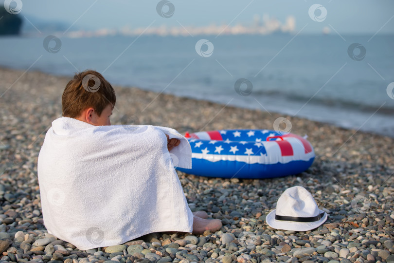 Скачать Мальчик, завернутый в полотенце, сидит на галечном пляже рядом с поплавком в цветах американского флага и в шляпе тихим утром на берегу моря. фотосток Ozero