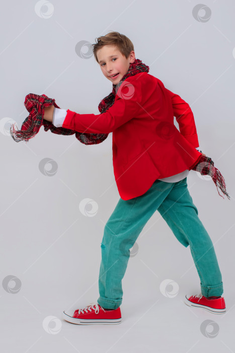 Скачать Забавный мальчик в красной куртке, зеленых джинсах и кроссовках в полный рост на сером фоне. Он делает большой шаг с поднятой ногой. Концепция - только вперед. фотосток Ozero