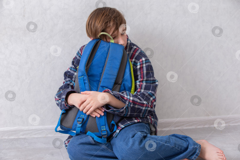 Скачать Ребенок сидит на полу, обнимая синий школьный рюкзак, в уютной комнате после обеда. Он устал от учебы. фотосток Ozero