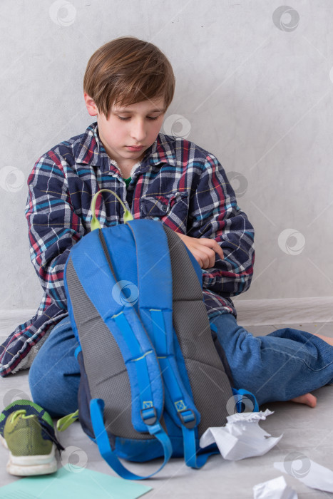 Скачать Мальчик сидит на полу и распаковывает школьные принадлежности из синего рюкзака в светлой комнате фотосток Ozero