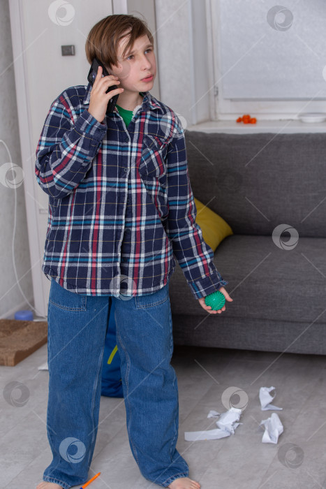 Скачать Мальчик в клетчатой рубашке разговаривает по телефону, стоя в грязной гостиной со смятой бумагой фотосток Ozero