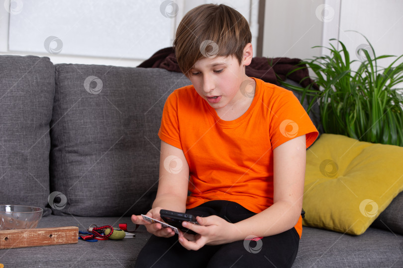 Скачать Мальчик в оранжевой рубашке пользуется смартфоном, сидя дома на сером диване фотосток Ozero