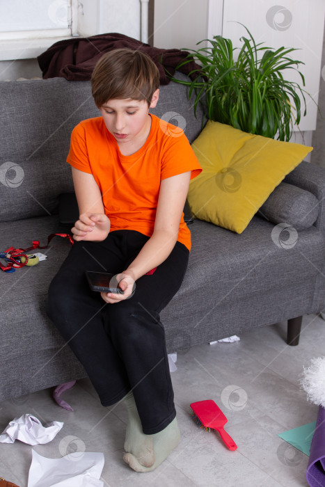 Скачать Мальчик в оранжевой рубашке сидит на диване и пользуется смартфоном в грязной гостиной фотосток Ozero