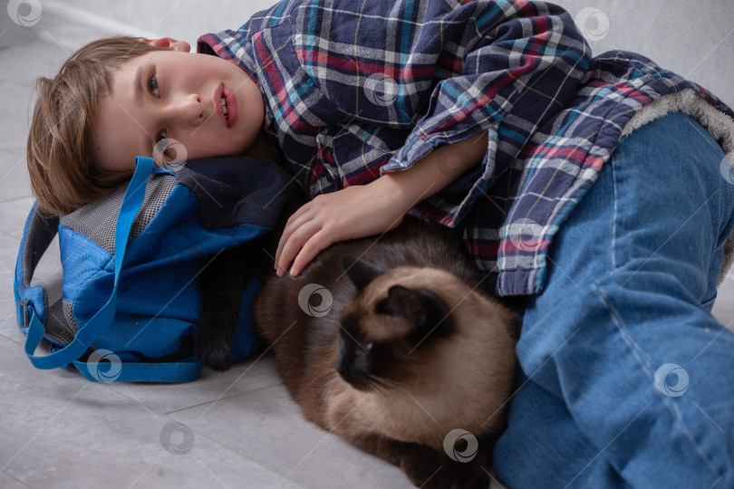 Скачать Маленький мальчик расслабляется на полу с синим рюкзаком в руках, обнимая коричневого кота в уютной домашней обстановке фотосток Ozero