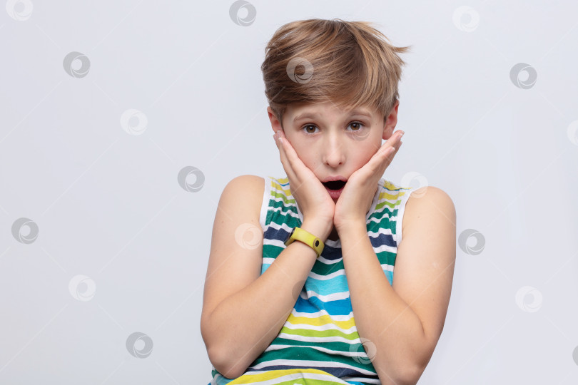 Скачать Маленький мальчик изображает удивление с широко раскрытыми глазами и прижатыми к щекам руками, одетый в яркую полосатую рубашку на простом фоне фотосток Ozero