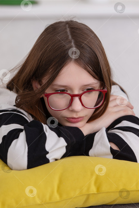 Скачать Молодая девушка в красных очках отдыхает дома днем на желтой подушке, погруженная в свои мысли фотосток Ozero