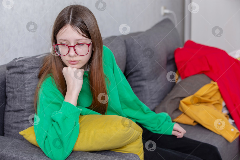 Скачать Девочка-подросток в зеленой толстовке сидит на сером диване с задумчивым выражением лица, вокруг разбросана разноцветная одежда фотосток Ozero
